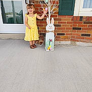 Madilynn is registered to the contest to win money with this photo: brick_wall, child, decoration, door, easter_bunny, eggs, footwear, girl, happy, holiday, outdoor, person, porch, shutter, sidewalk, smiling, spring, summer, window, yellow_dress