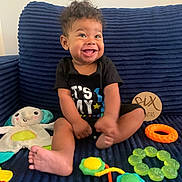 Haze is registered to the contest to win money with this photo: baby, child, smiling, curly_hair, black_shirt, toys, stuffed_animal, blue_couch, indoor, happy, sitting, foot, hand, colorful, playful, cute, infant, portrait, young_child, comfortable