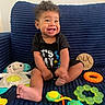 baby, child, smiling, curly_hair, black_shirt, toys, stuffed_animal, blue_couch, indoor, happy, sitting, foot, hand, colorful, playful, cute, infant, portrait, young_child, comfortable