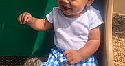 Haze is registered to the contest to win money with this photo: baby, child, smiling, curly_hair, white_shirt, blue_shorts, checkered_shorts, playground, slide, outdoor, sunlight, happy, sitting, person, park, wood_chips, daylight, cute, infant, play
