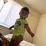 Haze is registered to the contest to win money with this photo: toddler, child, pacifier, green_onesie, toys, bed, indoor, lamp, window, curious, standing, person, room, baby, colorful, clothing, furniture, portrait, home, play