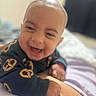 baby, child, infant, smiling, laughing, portrait, face, bald, indoor, bedroom, bed, blanket, clothing, black_shirt, patterned_clothing, sitting, happy, cozy, close_up, photograph