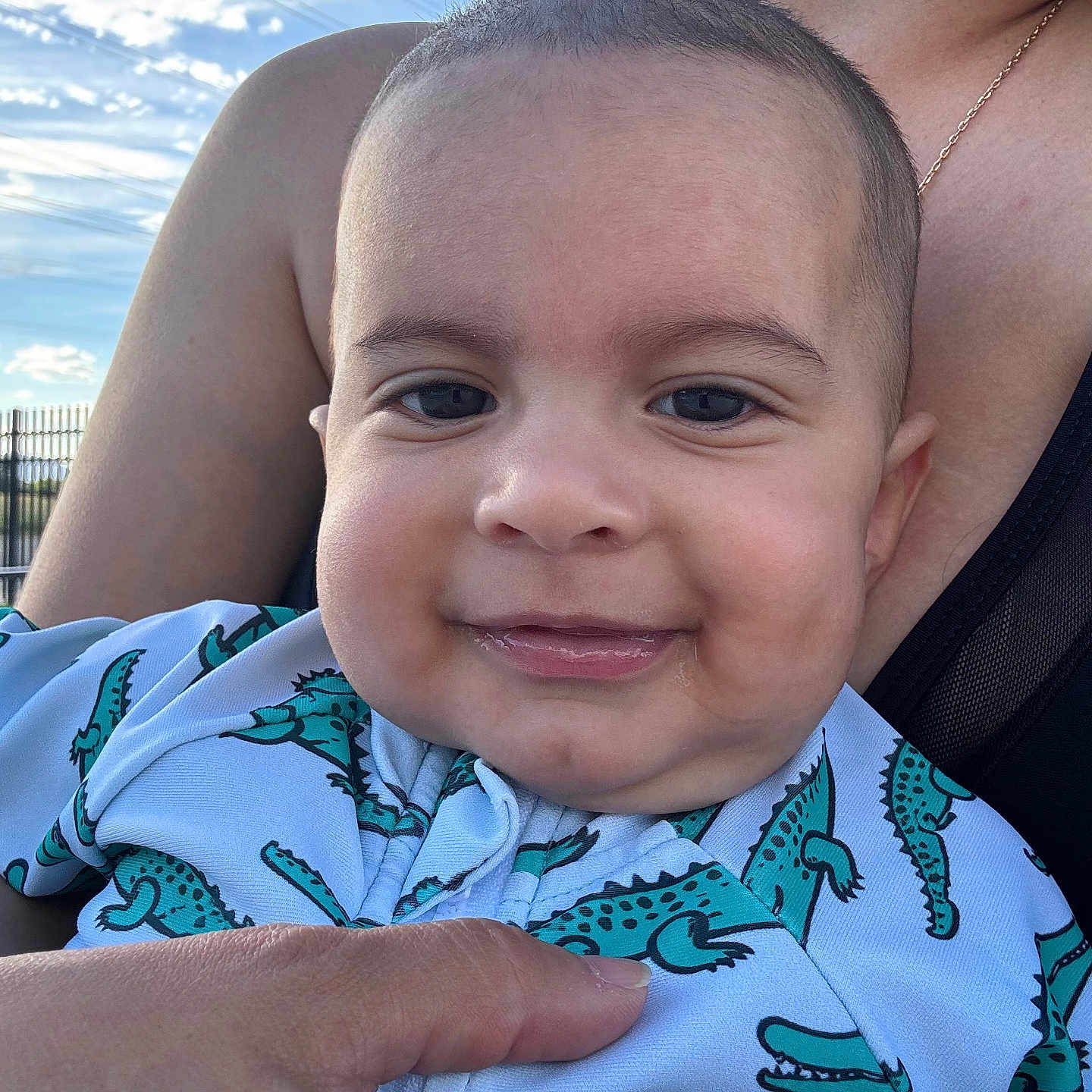 Thiago joined the competition — help win amazing prizes! baby, infant, child, smiling, portrait, close_up, face, person, adult, parent, cuddling, outdoor, sky, clouds, sunlight, blue_clothing, alligator_pattern, onesie, family, happy