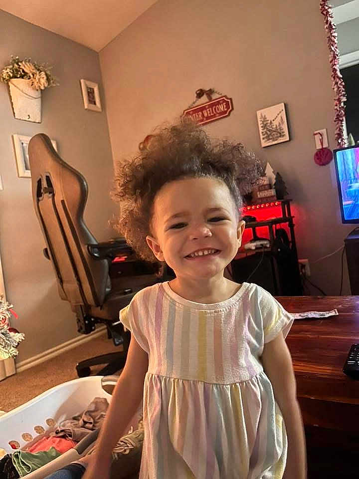 Harley is registered to the contest to win money with this photo: child, toddler, smiling, curly_hair, dress, indoor, living_room, furniture, chair, table, decorations, television, carpet, wooden_table, laundry_basket, home, person, happy, portrait, cute