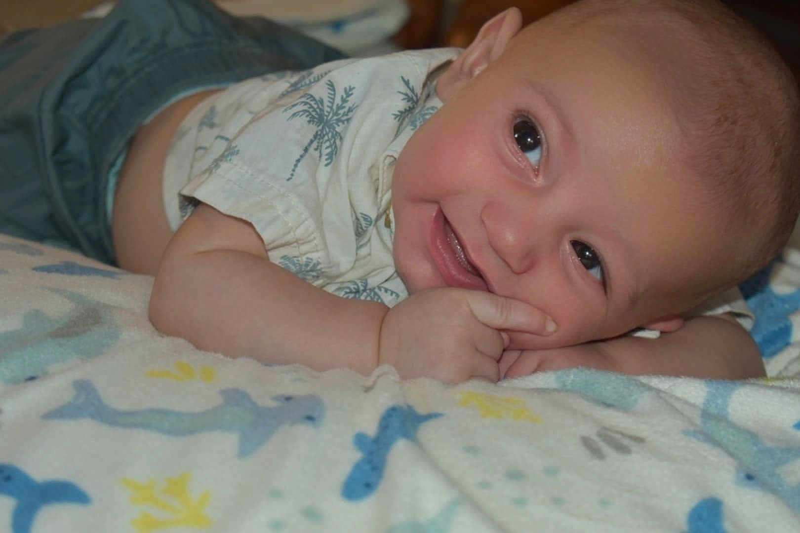 Apollo joined the competition — help win amazing prizes! baby, infant, smile, face, hand, blanket, pattern, blue, yellow, clothing, toddler, skin, cute, lying_down, cheeky, indoors, happy, closeup, person, child