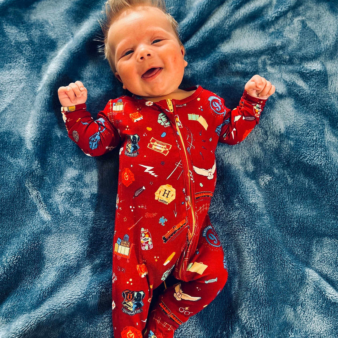 Henry is registered to the contest to win money with this photo: baby, child, smiling, onesie, red_clothing, blanket, blue_blanket, happy, infant, lying_down, cute, soft_texture, patterned_clothing, face, portrait, young_child, cozy, sleepwear, hands_up, indoors