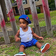 Dekhari joined the competition — help win amazing prizes! toddler, child, grass, wooden_fence, cap, blue_shorts, white_tank_top, sneakers, necklace, outdoor, residential_house, trees, greenery, casual_clothing, sitting, curious, daylight, fashion, young_child, sideways_cap