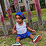 toddler, child, grass, wooden_fence, cap, blue_shorts, white_tank_top, sneakers, necklace, outdoor, residential_house, trees, greenery, casual_clothing, sitting, curious, daylight, fashion, young_child, sideways_cap