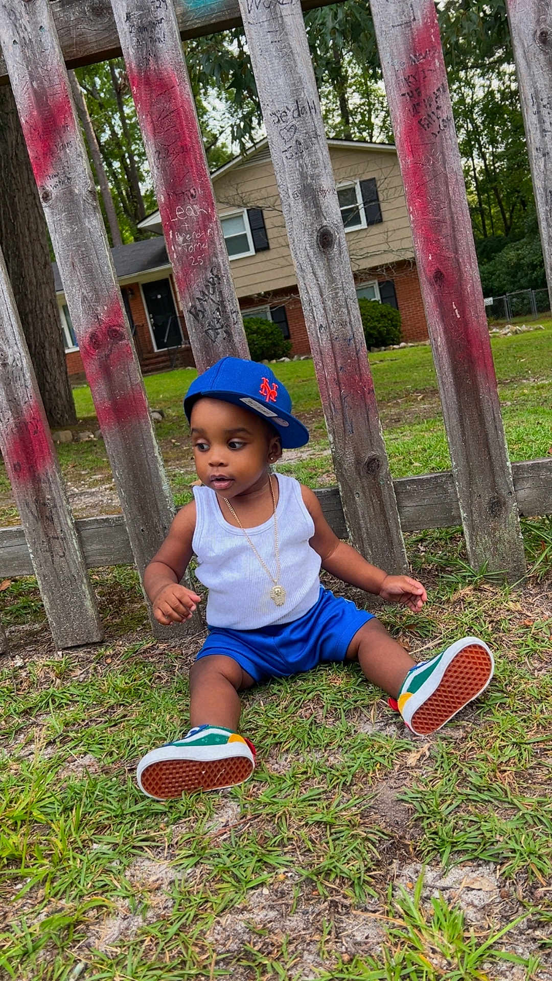 Dekhari joined the competition — help win amazing prizes! toddler, child, grass, wooden_fence, cap, blue_shorts, white_tank_top, sneakers, necklace, outdoor, residential_house, trees, greenery, casual_clothing, sitting, curious, daylight, fashion, young_child, sideways_cap