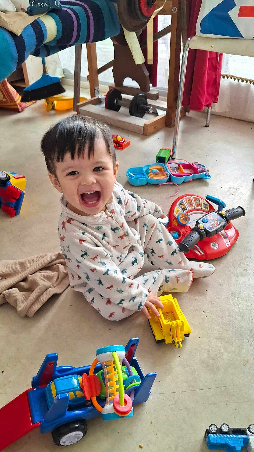 Ewen a rejoint le concours — aidez-le/la à gagner de superbes lots ! toddler, child, smiling, toy, toy_truck, floor, pajamas, animal_print, indoor, playtime, happy, colorful, plastic_toy, vehicle_toy, chair, blanket, curtain, window, person, casual