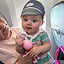 Isla-Rose is registered to the contest to win money with this photo: baby, child, adult, airplane_interior, window, toy, pink_toy, pilot_hat, striped_dress, holding, seated, curious, innocent, person, face, hand, clothing, travel, family, indoor