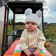 Isla-Rose joined the competition — help win amazing prizes! baby, child, hat, pom_poms, orange_socks, tractor, pumpkin_patch, outdoor, field, cloudy_sky, cute, serious_expression, knitwear, fall, autumn, greenery, farm, hands, face, sitting