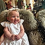 Mila is registered to the contest to win money with this photo: baby, infant, white_dress, headband, bow, fur_cushion, smiling, tongue_out, indoor, people, adults, blurred_background, table, chair, casual_clothing, warm_lighting, portrait, cute, happy, seated