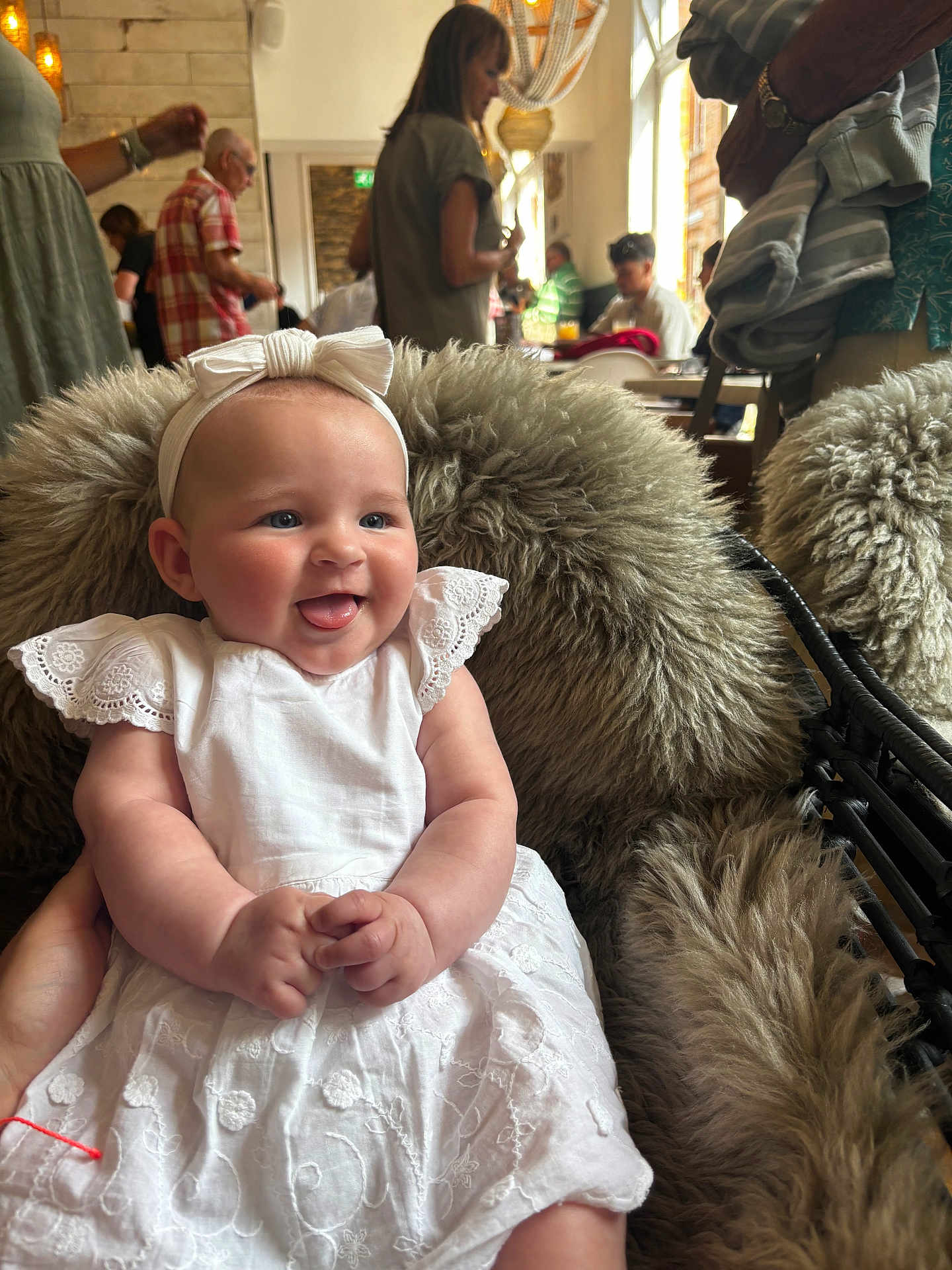 Mila is registered to the contest to win money with this photo: baby, infant, white_dress, headband, bow, fur_cushion, smiling, tongue_out, indoor, people, adults, blurred_background, table, chair, casual_clothing, warm_lighting, portrait, cute, happy, seated