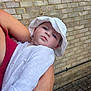 Mila joined the competition — help win amazing prizes! baby, child, white_hat, white_clothing, arm, adult, red_top, brick_wall, outdoor, pavement, person, holding, portrait, cute, face, skin, headwear, infant, tender_moment, casual