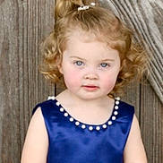 Harley is registered to the contest to win money with this photo: child, toddler, blue_dress, curly_hair, blonde_hair, portrait, outdoor, wooden_background, pearl_neckline, cute, innocent, girl, fashion, posing, smiling, person, young_child, natural_light, standing, casual