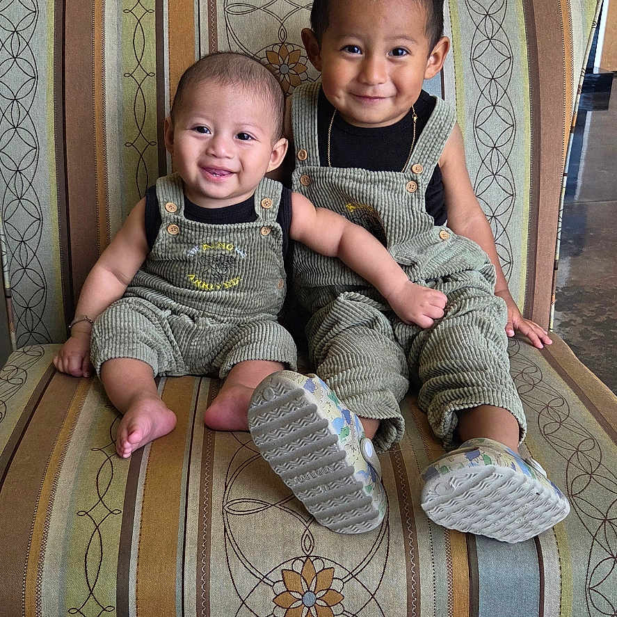 Milan joined the competition — help win amazing prizes! baby, barefoot, child, clothing, couch, cute, face, footwear, furniture, happy, head, indoor, overalls, patterned_fabric, person, portrait, siblings, sitting, smile, toddler