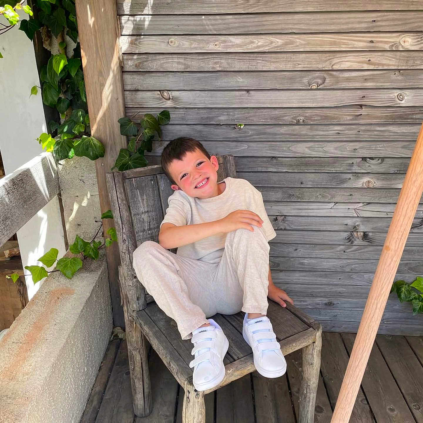 Barthélémy a rejoint le concours — aidez-le/la à gagner de superbes lots ! boy, casual_clothing, child, daylight, green_leaves, happy, ivy, natural_light, outdoor, person, plank_floor, plant, relaxed, rustic, seated, smile, white_shoes, wooden_chair, wooden_floor, wooden_wall