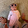 Riella is registered to the contest to win money with this photo: baby, pink_sunglasses, pink_outfit, carpet, hands, smiling, happy, child, indoor, person, cute, casual, accessory, watch, ring, feet, socks, toddler, playful, portrait