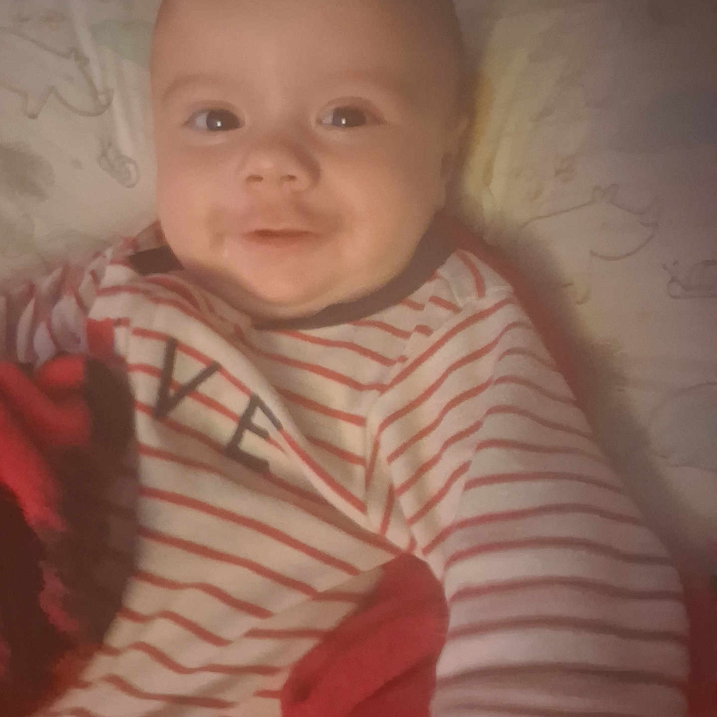 Noryan is registered to the contest to win money with this photo: baby, infant, child, smiling, portrait, striped_clothing, onesie, bed, blanket, pillow, stuffed_animal, red, black, indoor, cozy, lying_down, close_up, face, happy, photograph