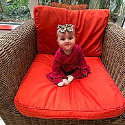 Aleena is registered to the contest to win money with this photo: baby, child, wicker_chair, orange_cushion, red_clothing, headband, bow, indoor, window, greenery, plant, smiling, cute, sitting, infant, portrait, floor, natural_light, home, decor