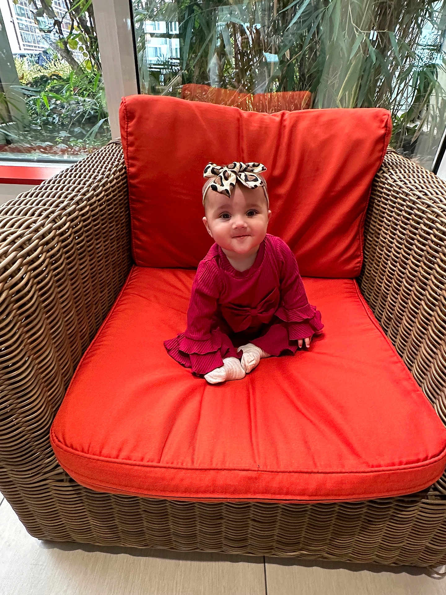 Aleena is registered to the contest to win money with this photo: baby, child, wicker_chair, orange_cushion, red_clothing, headband, bow, indoor, window, greenery, plant, smiling, cute, sitting, infant, portrait, floor, natural_light, home, decor