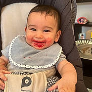 Isaac a rejoint le concours — aidez-le/la à gagner de superbes lots ! baby, child, smiling, bib, high_chair, messy_face, indoor, clothing, checkered_pants, happy, person, seatbelt, furniture, face, hand, foot, hair, table, plate, decor