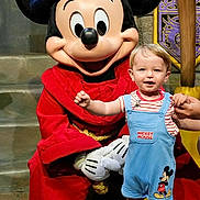 Jacob is registered to the contest to win money with this photo: child, toddler, mickey_mouse, costume, wizard_hat, red_clothing, blue_overalls, red_shoes, character, theme_park, stone_floor, stone_wall, purple_decor, gold_decor, holding_hands, smiling, portrait, indoor, person, fantasy