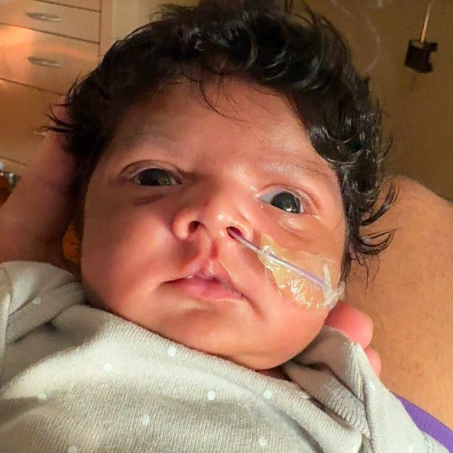Shay is registered to the contest to win money with this photo: adult_hand, baby, care, closeup, curly_hair, eyes, face, hand, healthcare, hospital, indoors, infant, lying_down, medical_equipment, nasal_tube, newborn, patient, person, polka_dot_clothing, skin