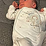 newborn, baby, infant, lying_down, white_clothing, bottle, blanket, gray_surface, embroidered_text, 2025, cozy, soft_texture, star_pattern, cute, small_hand, facial_expression, indoors, resting, sleepwear, baby_garment