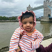 Kunj joined the competition — help win amazing prizes! baby, infant, child, striped_clothing, bonnet, person, portrait, outdoor, river, bridge, tower_bridge, landmark, sky, cloudy, hand, holding, water, urban, tourist_spot, casual_clothing