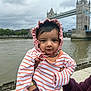 baby, infant, child, striped_clothing, bonnet, person, portrait, outdoor, river, bridge, tower_bridge, landmark, sky, cloudy, hand, holding, water, urban, tourist_spot, casual_clothing