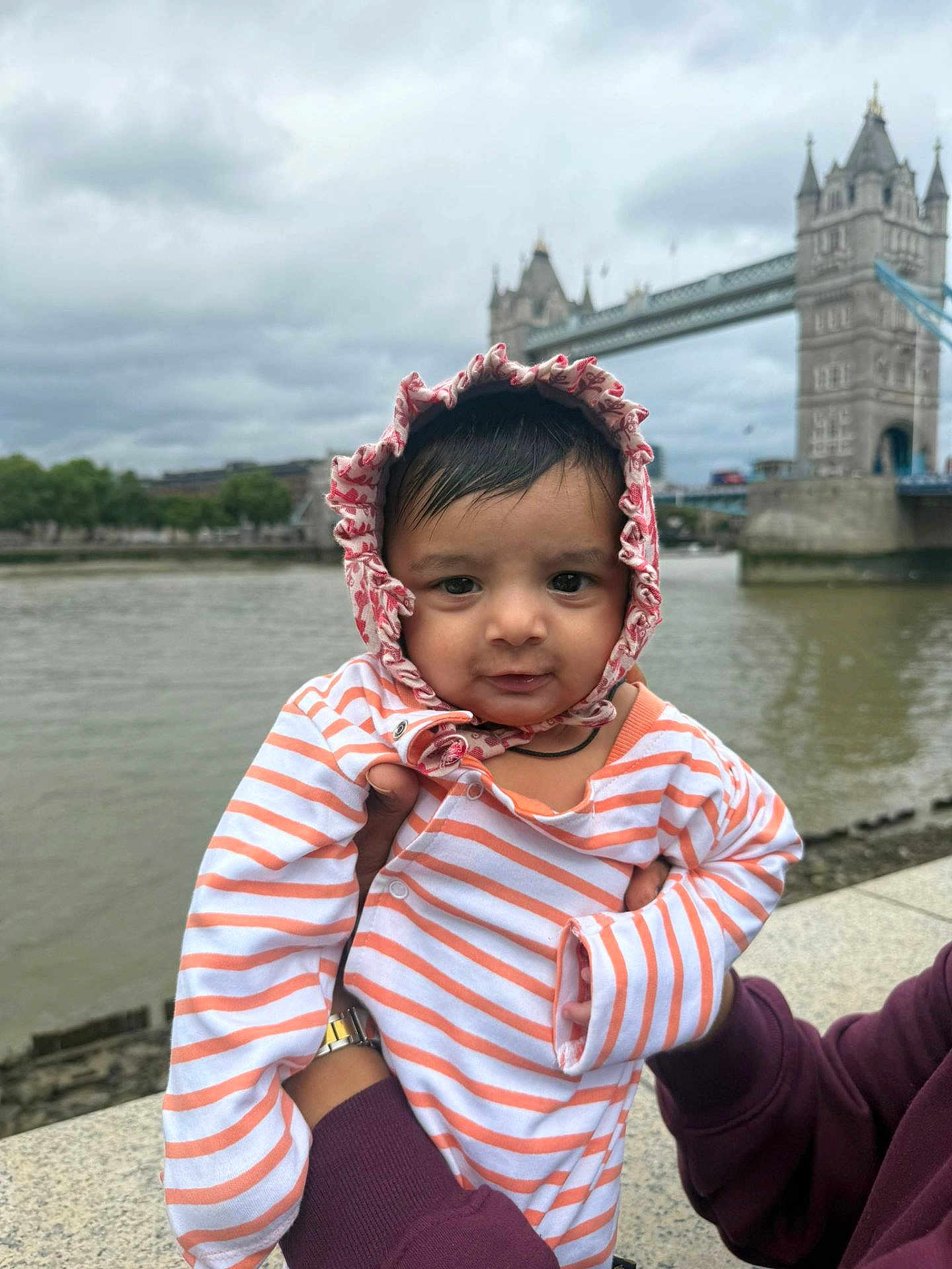 Kunj joined the competition — help win amazing prizes! baby, infant, child, striped_clothing, bonnet, person, portrait, outdoor, river, bridge, tower_bridge, landmark, sky, cloudy, hand, holding, water, urban, tourist_spot, casual_clothing