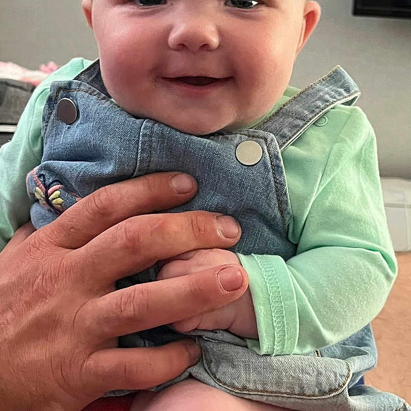 Remi joined the competition — help win amazing prizes! adult_hand, baby, bow, child, chubby_cheeks, clothing, cute, denim_overalls, face, hand, happy, headband, indoor, infant, person, portrait, sitting, skin, smiling, young