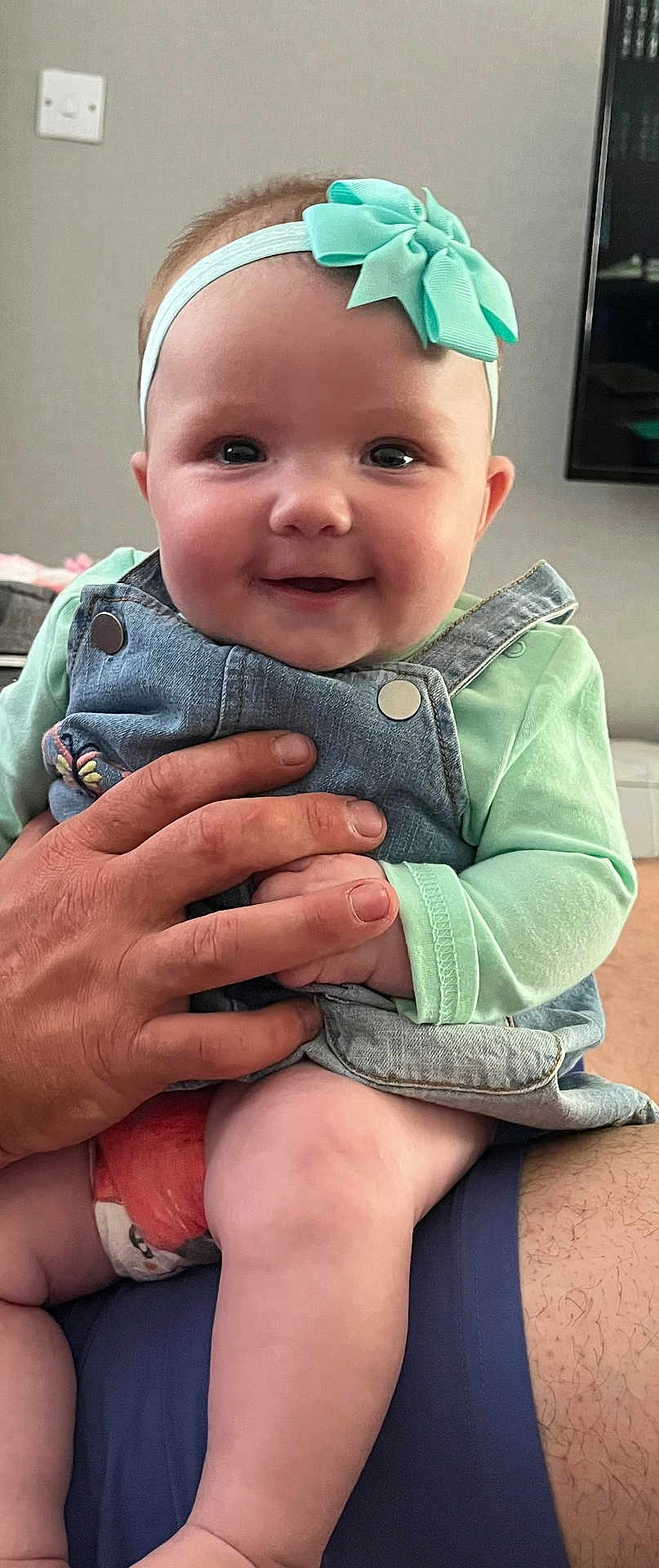 Remi joined the competition — help win amazing prizes! baby, child, smiling, headband, bow, denim_overalls, hand, adult_hand, indoor, skin, face, cute, chubby_cheeks, sitting, clothing, person, infant, happy, portrait, young