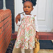Gemma is registered to the contest to win money with this photo: toddler, child, curly_hair, bow, dress, paisley_pattern, sunglasses, handbag, white_long_sleeve, tights, shoes, doorstep, brick_wall, doormat, outdoor, standing, person, fashion, cute, portrait