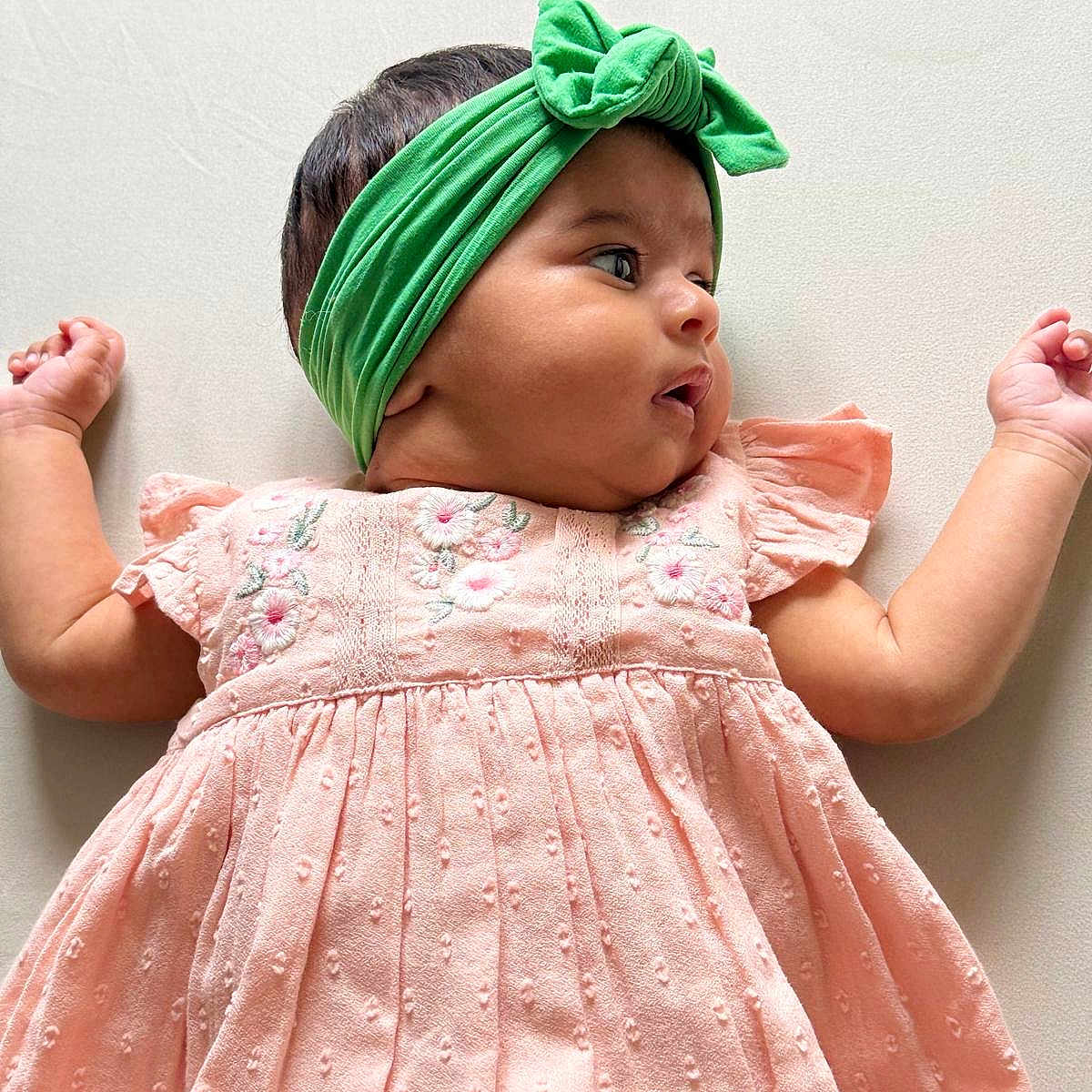 Elleen is registered to the contest to win money with this photo: arms_outstretched, baby, child, closeup, clothing, curious, cute, embroidery, fabric, green, headband, infant, laying_down, person, pink_dress, portrait, side_profile, skin, soft_background, young_child
