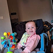 Rosie-Mae is registered to the contest to win money with this photo: baby, child, smiling, pink_clothing, bib, toy, high_chair, indoor, face, happy, seat, furniture, colorful, play, person, infant, cheerful, young_child, home, accessory