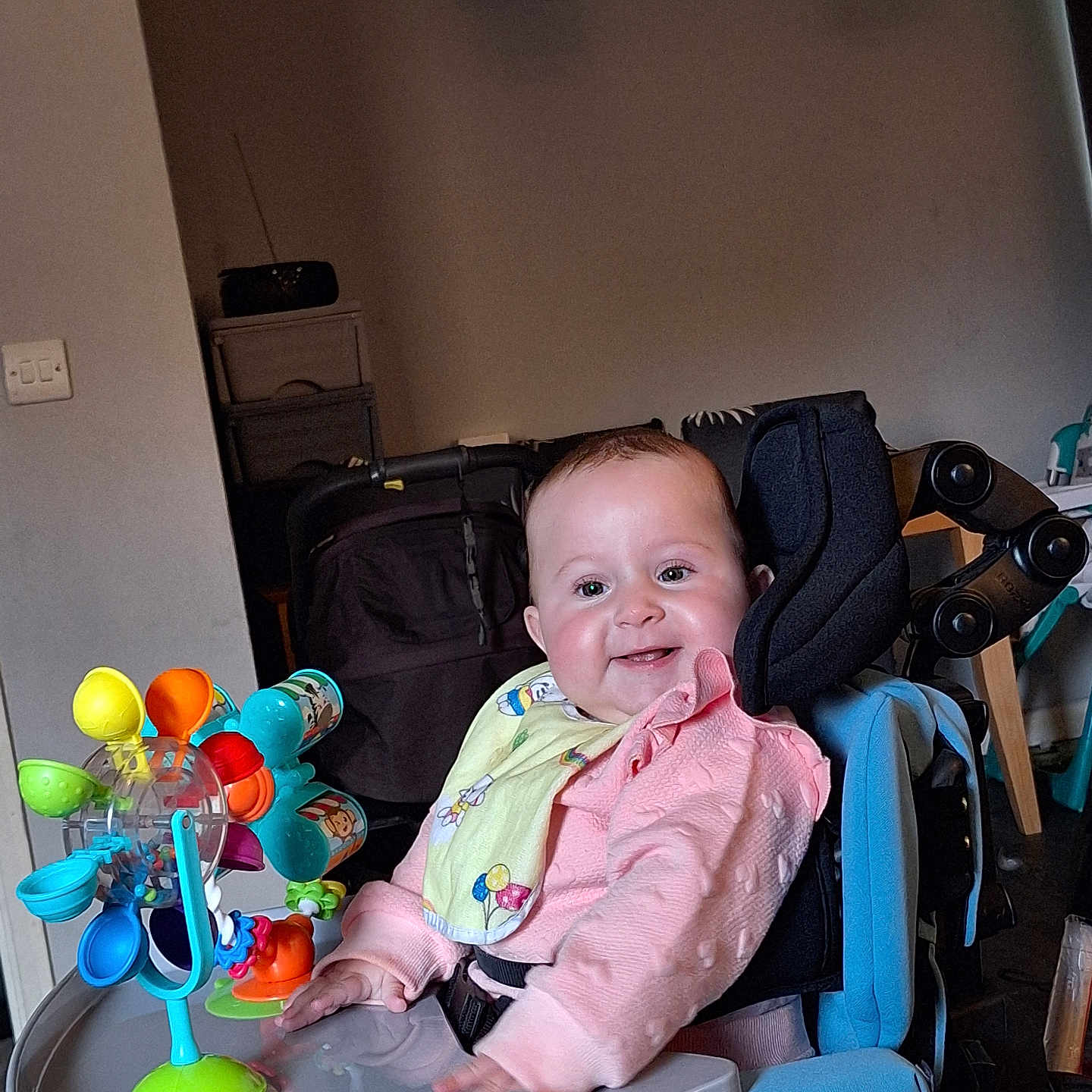 Rosie-Mae is registered to the contest to win money with this photo: accessory, baby, bib, cheerful, child, colorful, face, furniture, happy, high_chair, home, indoor, infant, person, pink_clothing, play, seat, smiling, toy, young_child