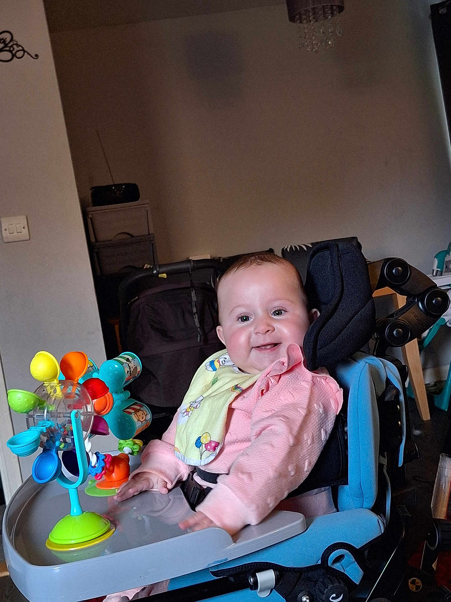 Rosie-Mae is registered to the contest to win money with this photo: baby, child, smiling, pink_clothing, bib, toy, high_chair, indoor, face, happy, seat, furniture, colorful, play, person, infant, cheerful, young_child, home, accessory