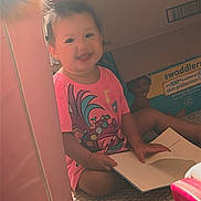 Harley is registered to the contest to win money with this photo: toddler, child, smiling, pink_shirt, book, carpet, box, homestyle_seasoned_croutons, swaddlers_box, indoor, play_area, sitting, happy, person, cute, young_child, floor, reading, toy_storage, casual_clothing