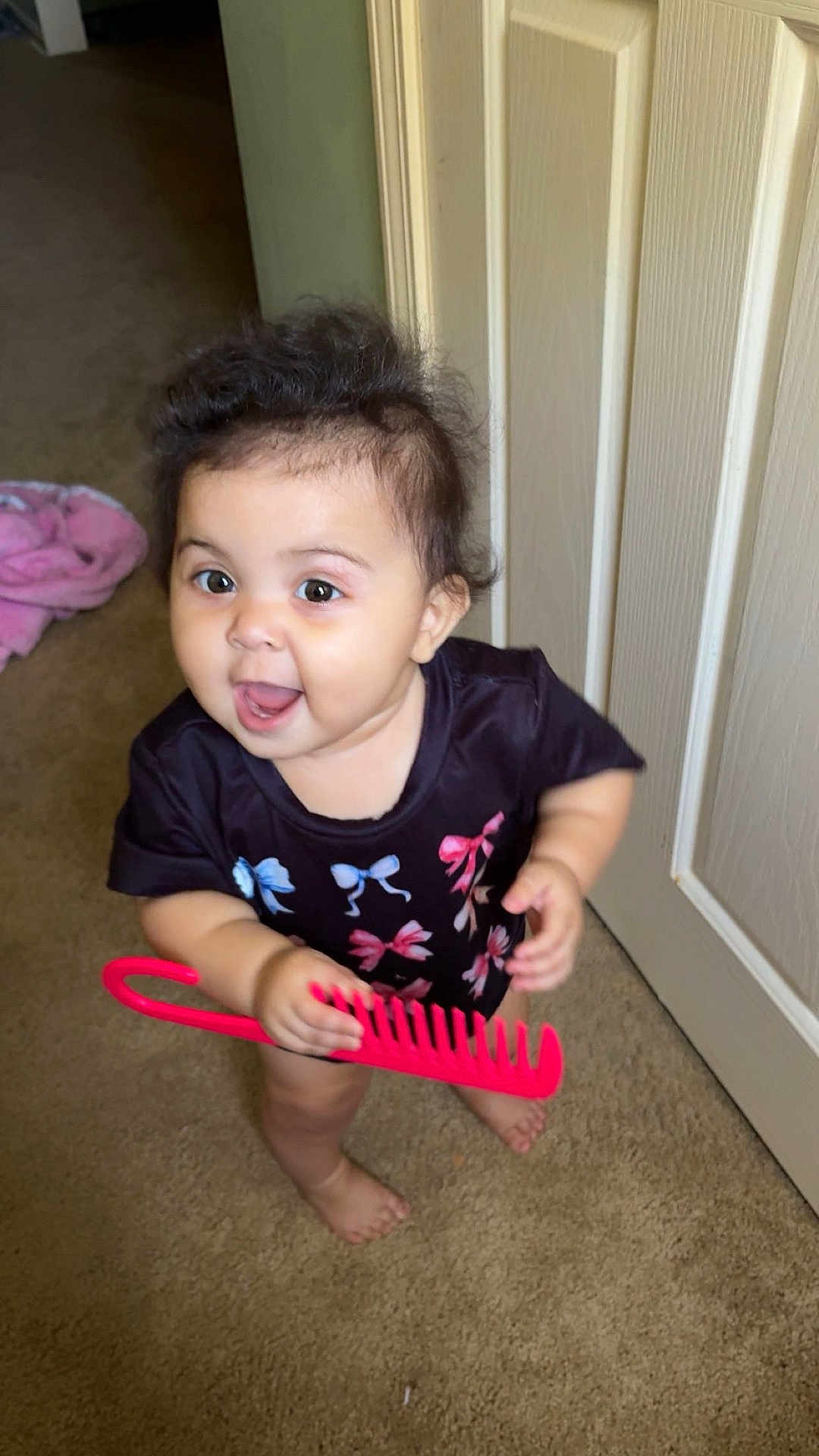 Nova joined the competition — help win amazing prizes! baby, child, barefoot, carpet, comb, door, floor, hair, indoors, person, shirt, smiling, standing, toy, young_child, playful, happy, colorful_clothing, pink, black_shirt