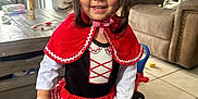 Hazelyn is registered to the contest to win money with this photo: toddler, child, smiling, costume, red_hood, indoor, living_room, toy, sofa, table, curtain, floor_tile, happy, girl, dress, cape, playtime, cute, person, portrait