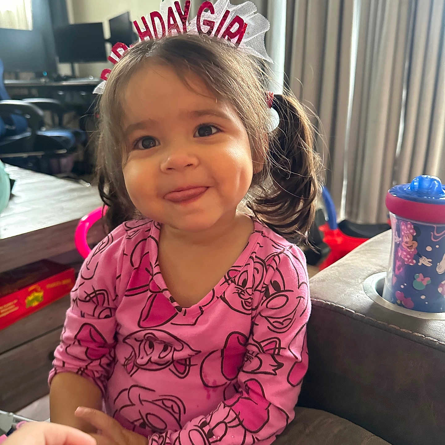 Hazelyn is registered to the contest to win money with this photo: birthday_headband, cartoon_print, chair, child, couch, curtains, cute, dimples, furniture, girl, hand, indoor, living_room, pigtails, pink_clothing, sippy_cup, smiling, table, toddler, tongue_out