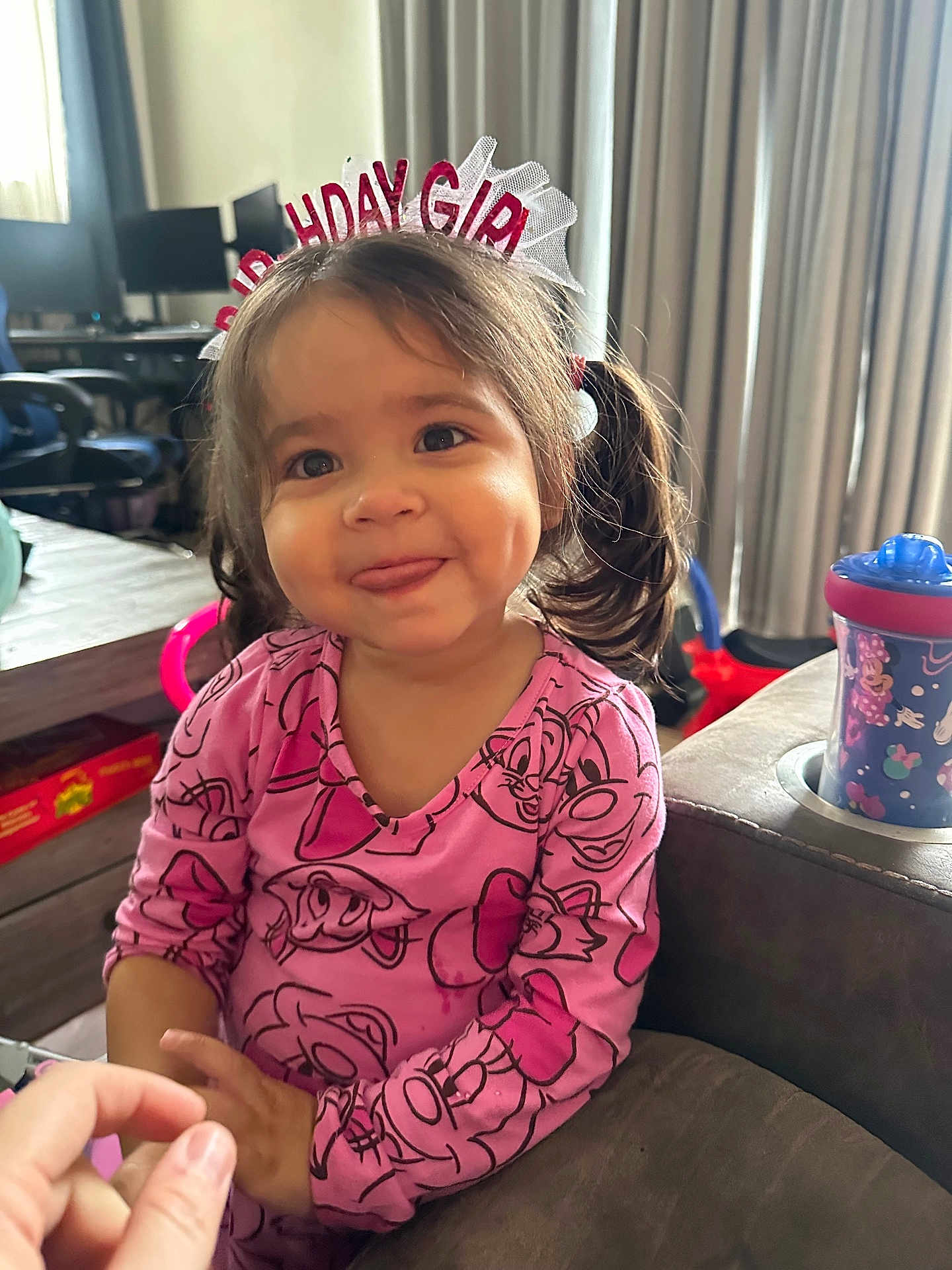 Hazelyn is registered to the contest to win money with this photo: toddler, girl, birthday_headband, pink_clothing, dimples, tongue_out, indoor, couch, sippy_cup, curtains, table, chair, cartoon_print, child, smiling, pigtails, hand, furniture, living_room, cute