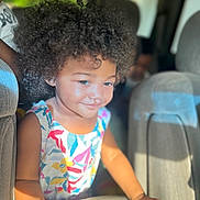 Dakota is registered to the contest to win money with this photo: child, toddler, curly_hair, afro, smiling, colorful_dress, floral_pattern, sunlight, car_interior, seat, hand, portrait, happy, cute, person, indoors, daylight, skin, young_child, people