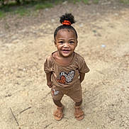 Dakota is registered to the contest to win money with this photo: child, toddler, smiling, curly_hair, orange_scrunchie, brown_outfit, heart_pattern, sand, outdoor, footwear, sandal, happy, standing, person, cute, young_child, daylight, nature, greenery, playful