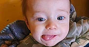 Jasper is registered to the contest to win money with this photo: baby, child, infant, face, portrait, wide_eyes, expression, smiling, tongue, camouflage_clothing, blanket, bed, cozy, indoors, orange_wall, brown_hair, head, sitting, close_up, cute