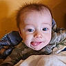 baby, child, infant, face, portrait, wide_eyes, expression, smiling, tongue, camouflage_clothing, blanket, bed, cozy, indoors, orange_wall, brown_hair, head, sitting, close_up, cute