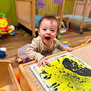 Jasper is registered to the contest to win money with this photo: baby, child, nursery, crib, toys, indoor, portrait, smiling, laughing, happy, sitting, standing, artwork, painting, yellow, black, wooden_furniture, playroom, infant, home